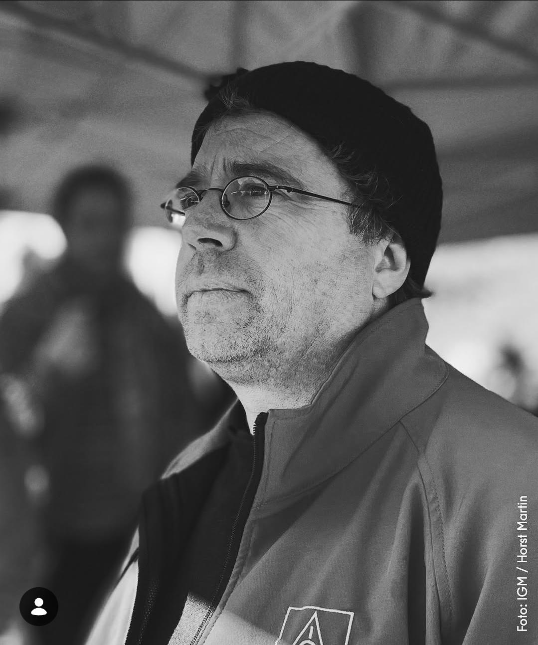 Ein Schwarz-Weiß-Foto von Christoph Ellinghaus in einer Nahaufnahme. Er trägt eine dunkle Strickmütze und eine Brille mit dünnen, runden Bügeln. Sein Blick ist nach links gerichtet und sein Ausdruck wirkt nachdenklich. Er trägt eine Jacke mit einem Logo auf der Brust. Im unscharfen Hintergrund ist eine Zeltstruktur und eine weitere Person zu erkennen.